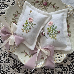 Op de afbeelding: Twee rechthoekige speldenkussens van witte stof met geborduurde roze rozentakjes met groene bladeren. Elk kussentje is versierd met een decoratieve strik van mauve en crèmekleurig lint. Eén kussentje heeft een parelknopspeld en een bloemknopspeld.
