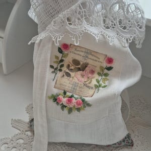 Könnte beinhalten: Weiße Leinentasche mit Spitzenbesatz und dekorativem Blumendesign. Die Tasche zeigt ein gesticktes Bild von rosa Rosen und den Text "PARIS". Ein langer weißer Riemen hängt von der Tasche.