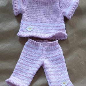 May include: A pink and white striped knitted doll outfit with a daisy applique. The top has short sleeves and a scalloped neckline. The trousers have a wide leg and a scalloped hem.
