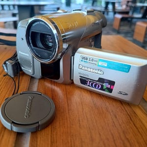 May include: A silver Panasonic NV-GS75 camcorder with a 3CCD lens system and a flip-out LCD screen. The screen displays text including USB 2.0, Panasonic, and 3CCD. A separate circular Panasonic lens cap sits in front of the device on the surface.