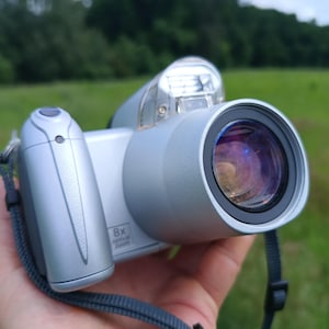 May include: A silver digital camera featuring a prominent lens with a dark glass element, a pop-up flash unit on top, and a textured grip area. The camera body displays the text 8x optical zoom near the lens barrel and includes a grey wrist strap.
