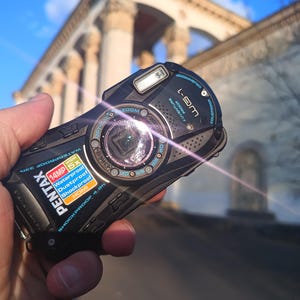 以下が含まれることがあります： 黒いPentax WG-1デジタルカメラ。頑丈なデザインで青いアクセントが特徴です。前面には14MP、5倍光学ズーム、防水、防塵、耐衝撃、HDMIの文字が記載されています。レンズは中央に配置され、リングライトが備わっています。