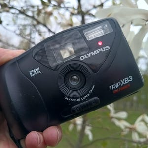 Könnte beinhalten: Schwarze Olympus Trip XB3 Kamera mit 34mm Objektiv. Die Kamera hat einen eingebauten Blitz und der Text "OLYMPUS TRIP XB3 BIG FINGER" ist sichtbar. Die Kamera wird in einer Hand vor einem Hintergrund aus weißen Blüten gehalten.
