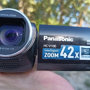 Op de afbeelding: Een zwarte Panasonic HC-V100 camcorder met een lens gemarkeerd met Optical Zoom f=2.38-81.0mm 1:1.8. Het zijpaneel bevat tekst met Full HD 1920x1080, Intelligent Zoom 42x, 32.5mm Wide, Power O.I.S., AVCHD en een SD XC geheugenkaartlogo.