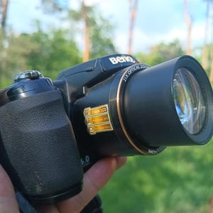 May include: A black BenQ digital camera featuring a 21x optical zoom lens with a copper-colored ring. The camera body has a textured grip and a sticker on the lens barrel displaying technical specifications including 21x zoom and LCD details.
