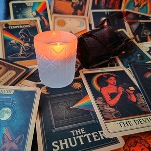 Op de afbeelding: Een matglazen kaarshouder met een geometrisch ruitpatroon gloeit met een warm oranje licht, omringd door een verzameling tarotkaarten met camera-thema illustraties, waaronder kaarten met de tekst The Shutter en The Devil.