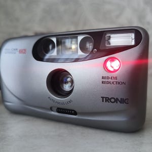 May include: Silver Tronic point-and-shoot film camera with a red light indicating red-eye reduction. The camera has an auto-focus lens and the text "CAM KK35" is visible. A black strap is attached.