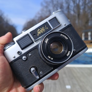 May include: A silver and black FED 4 camera with a black lens. The camera has a black leather grip and a silver metal body. The lens has a black metal ring with the numbers 2137327 and 24/35 printed on it.