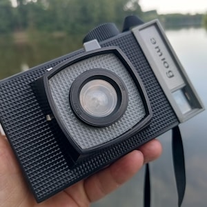 May include: A black plastic film camera with a textured grid pattern on the body and a square lens housing. The camera features a silver rectangular plate on the top right with the Cyrillic text "элиос" printed in black, and a black strap.