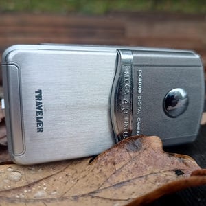 May include: A silver Traveler DC4000 digital camera with a brushed metal finish. The device features a prominent chrome-colored accent strip, a circular lens cover, and text reading "TRAVELER" and "DC4000 DIGITAL CAMERA MEGA 4.0 PIXELS."
