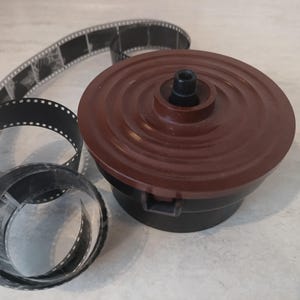 Może przedstawiać: Browning plastikowa szpula do wywoływania filmu z owiniętym wokół nią czarnym paskiem filmu.
