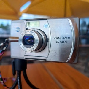 May include: A silver Minolta DiMAGE G600 digital camera featuring a GT Hexanon 8-24mm 1:2.8-4.9 lens. The camera body has a metallic finish with the model name printed on the front and a colorful patterned wrist strap attached to the side.