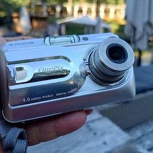 May include: A silver Fujifilm FinePix A340 digital camera featuring a 4.0 mega pixels lens, a built-in flash, and a gray wrist strap. The front of the camera displays the brand name, model name, and technical specifications in black text.