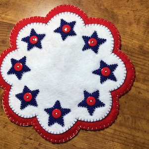 Puede incluir: Un posavasos patri&oacute;tico de fieltro con borde festoneado. Presenta un centro blanco, un borde rojo y estrellas azules, cada una con un bot&oacute;n rojo en el centro. El dise&ntilde;o est&aacute; cosido con hilo blanco.