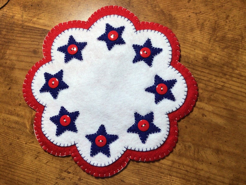 Puede incluir: Un posavasos patri&oacute;tico de fieltro con borde festoneado. Presenta un centro blanco, un borde rojo y estrellas azules, cada una con un bot&oacute;n rojo en el centro. El dise&ntilde;o est&aacute; cosido con hilo blanco.