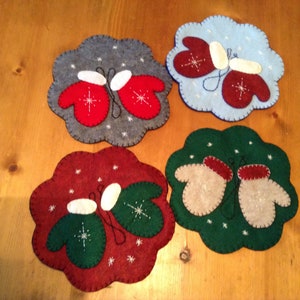 May include: Four felt ornaments shaped like mittens with snowflakes on a wooden surface. The mittens are in pairs, with each pair a different color: red, green, gray, and beige.