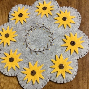May include: A gray felt table runner with a brown stitched border and eight yellow felt sunflowers with brown button centers. The sunflowers are arranged in a circle around a brown stitched wreath.