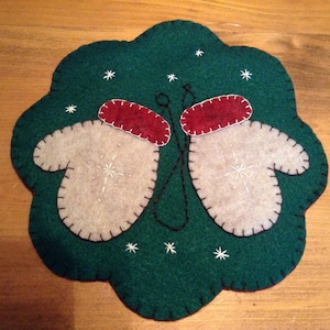 K&ouml;nnte beinhalten: Ein Filz-Weihnachtsornament mit gr&uuml;nem Hintergrund und zwei wei&szlig;en F&auml;ustlingen mit roten B&uuml;ndchen. Die F&auml;ustlinge sind mit schwarzem Faden zusammengen&auml;ht. Auf dem gr&uuml;nen Hintergrund sind wei&szlig;e Schneeflocken gestickt.
