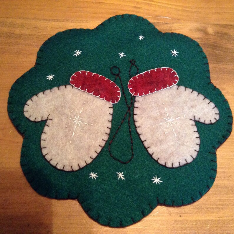 K&ouml;nnte beinhalten: Ein Filz-Weihnachtsornament mit gr&uuml;nem Hintergrund und zwei wei&szlig;en F&auml;ustlingen mit roten B&uuml;ndchen. Die F&auml;ustlinge sind mit schwarzem Faden zusammengen&auml;ht. Auf dem gr&uuml;nen Hintergrund sind wei&szlig;e Schneeflocken gestickt.