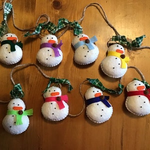 May include: A garland of handmade snowman ornaments. Each snowman is white with a colorful scarf, a carrot nose, and button eyes. They are strung together with twine and green plaid ribbon.