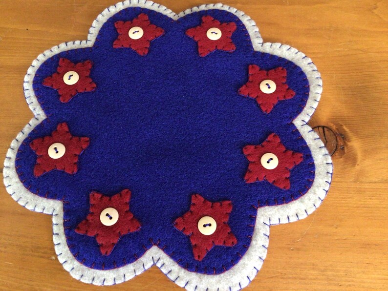 Puede incluir: Un posavasos de fieltro azul con borde festoneado, adornado con siete aplicaciones de estrellas rojas. Cada estrella tiene un bot&oacute;n blanco en el centro. El borde es blanco con un borde cosido. Un art&iacute;culo decorativo.