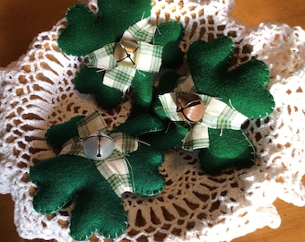 Puede incluir: Tres adornos de trébol de fieltro verde con campanas. Cada trébol está decorado con una cinta a cuadros y una campana. Los adornos están sobre un encaje blanco de ganchillo.