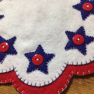 Puede incluir: Un mantel individual patri&oacute;tico de fieltro con borde festoneado. El mantel individual presenta fieltro blanco con aplicaciones de estrellas azules, cada una adornada con un bot&oacute;n rojo. El borde est&aacute; adornado con fieltro rojo, creando un aspecto festivo y artesanal.