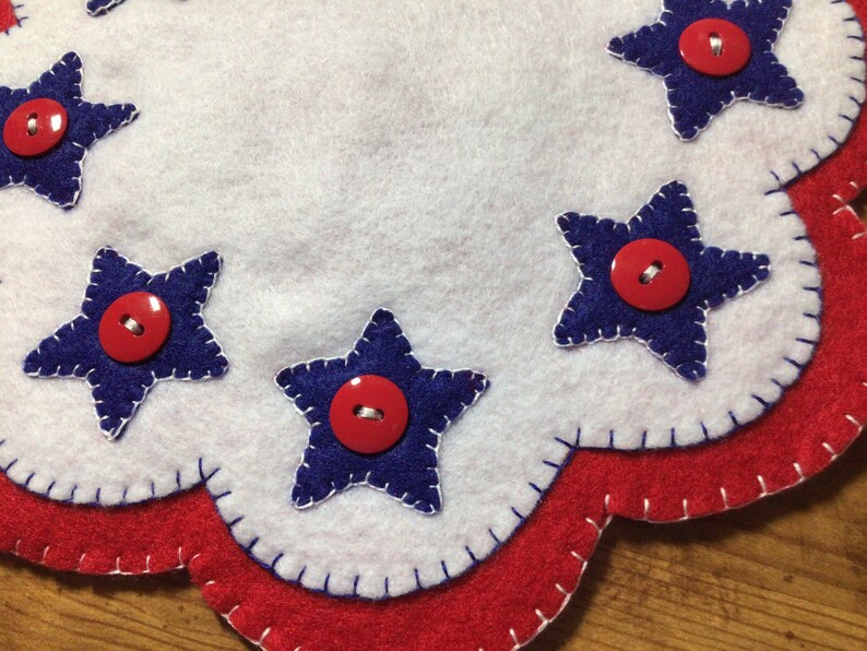 Puede incluir: Un mantel individual patri&oacute;tico de fieltro con borde festoneado. El mantel individual presenta fieltro blanco con aplicaciones de estrellas azules, cada una adornada con un bot&oacute;n rojo. El borde est&aacute; adornado con fieltro rojo, creando un aspecto festivo y artesanal.