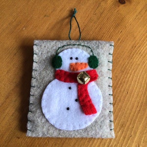 May include: A felt snowman ornament with a red scarf, green earmuffs, and a gold bell. The ornament is beige with white stitching around the edges.