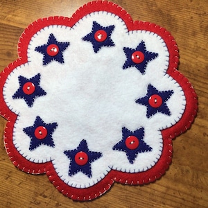 Puede incluir: Un posavasos patri&oacute;tico de fieltro hecho a mano con un centro blanco y un borde rojo festoneado. Siete estrellas azul marino, cada una con un bot&oacute;n rojo en el centro, est&aacute;n espaciadas uniformemente alrededor del centro blanco. El posavasos tiene un borde cosido.