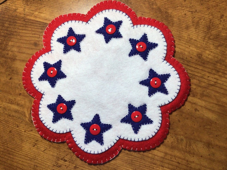 Puede incluir: Un posavasos patri&oacute;tico de fieltro hecho a mano con un centro blanco y un borde rojo festoneado. Siete estrellas azul marino, cada una con un bot&oacute;n rojo en el centro, est&aacute;n espaciadas uniformemente alrededor del centro blanco. El posavasos tiene un borde cosido.