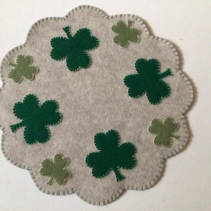 May include: A light gray felt placemat with a scalloped edge, decorated with green and light green shamrocks. The shamrocks are stitched onto the placemat, creating a festive St. Patrick's Day theme.