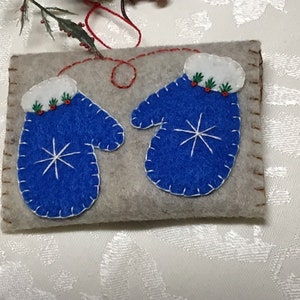 Könnte beinhalten: Ein Paar blaue Filzhandschuhe mit weißem Besatz und Schneeflocken, befestigt auf einem grauen Filzgrund. Die Handschuhe haben weiße Filzmanschetten und rote Filz-Stechpalmenbeeren.
