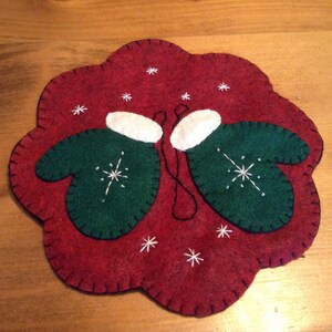 K&ouml;nnte beinhalten: Ein roter Filz-Anh&auml;nger mit zwei gr&uuml;nen Filz-Handschuhen mit wei&szlig;en Manschetten und Schneeflocken-Stickerei. Der Anh&auml;nger ist mit wei&szlig;en Schneeflocken-Stickereien verziert.