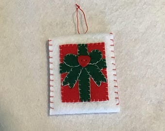 Puede incluir: Un adorno rectangular de fieltro con un borde blanco y costuras de festón rojas. El centro muestra un rectángulo de fieltro rojo con un lazo de fieltro verde oscuro y un pequeño botón rojo en forma de corazón. Un hilo rojo fino está unido arriba.