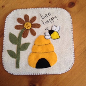 May include: A white felt square with a brown flower, a yellow bee, and a yellow beehive. The text "bee happy" is embroidered in brown thread.