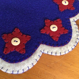 Puede incluir: Un posavasos de fieltro hecho a mano con base azul, ribete blanco y detalles de estrellas rojas. Cada estrella est&aacute; adornada con un bot&oacute;n blanco. El posavasos tiene un borde festoneado.
