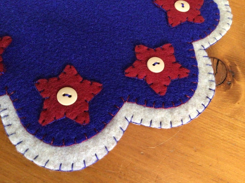 Puede incluir: Un posavasos de fieltro hecho a mano con base azul, ribete blanco y detalles de estrellas rojas. Cada estrella est&aacute; adornada con un bot&oacute;n blanco. El posavasos tiene un borde festoneado.
