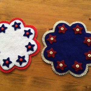 Puede incluir: Dos posavasos de fieltro con forma de flor. Uno es blanco con borde rojo y detalles de estrellas azules. El otro es azul con borde blanco y detalles de estrellas rojas. Cada estrella tiene un bot&oacute;n en el centro.