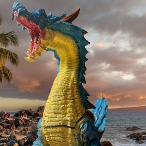 May include: A blue and yellow dragon figurine with an open mouth, standing on a rocky beach with a palm tree in the background.