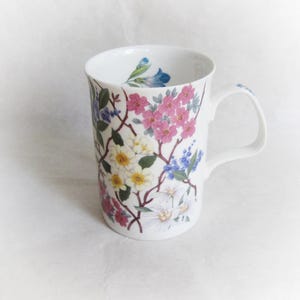 May include: White ceramic mug with a floral design featuring pink, white, and blue flowers. The mug has a handle and is approximately 4 inches tall.