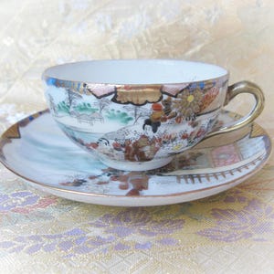 Puede incluir: Un juego de taza y platillo de porcelana vintage. La taza y el platillo presentan una escena pintada a mano con figuras, árboles y edificios en tonos marrones, verdes y dorados. La taza tiene un asa y un borde dorados. El platillo tiene un diseño a juego.