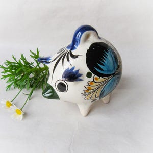 Può includere: Un salvadanaio di ceramica bianca con disegni floreali blu, gialli e neri. Il salvadanaio ha un piccolo foro in cima e un muso con un dettaglio dipinto di verde.