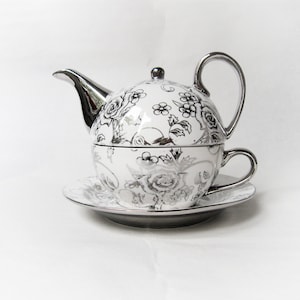 May include: A white ceramic tea set for one, featuring a teapot stacked on a teacup and saucer. The set is decorated with a metallic silver floral pattern. The teapot has a silver spout and handle, while the cup has a silver handle and rim.