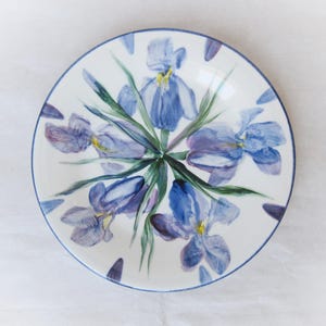 Pode incluir: Prato redondo de cerâmica branca com um desenho pintado à mão de flores de íris azuis e roxas e folhas verdes. O prato tem uma borda azul e está sobre um fundo branco. Um acento floral decorativo para a casa.