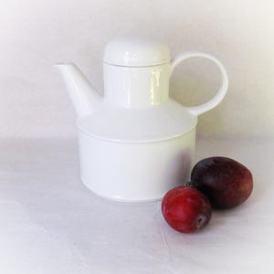 Könnte beinhalten: Eine weiße Keramik-Teekanne mit Deckel und gebogenem Griff. Zwei Pflaumen, eine rote und eine dunkelviolette, liegen neben der Teekanne. Die Teekanne hat einen zylindrischen Boden und einen Ausguss.