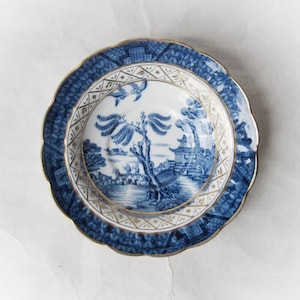 May include: A blue and white porcelain plate with a scalloped edge and gold trim. The plate features a detailed landscape scene in the center and a geometric pattern around the rim. The plate is a decorative item.