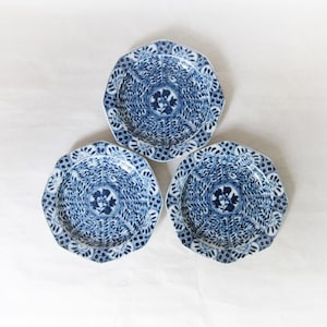 Könnte beinhalten: Drei blau-weiße Zierteller mit floralem Design. Die Teller haben einen gewellten Rand und ein detailliertes Muster. Die Teller sind auf einer weißen Oberfläche angeordnet.