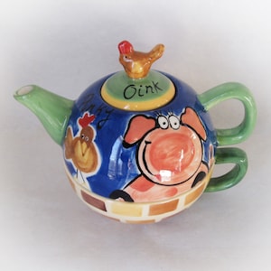 Peut inclure: Une théière bleue et verte avec un motif de cochon et de poulet. Le couvercle a un petit poulet sur le dessus et dit "Oink". La théière a une poignée de chaque côté.