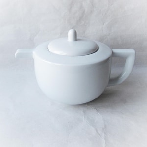 Può includere: Zuccheriera in ceramica bianca con coperchio e manico. La ciotola ha una forma arrotondata con un piccolo manico laterale e un coperchio con un piccolo pomello. La zuccheriera è su sfondo bianco.